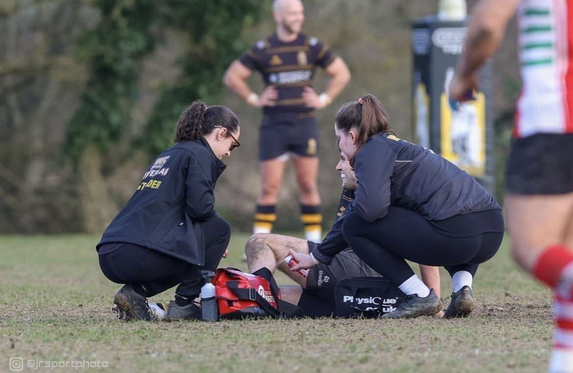 Two pitch-side first aiders kneeling to assess a rugby player’s leg during a match