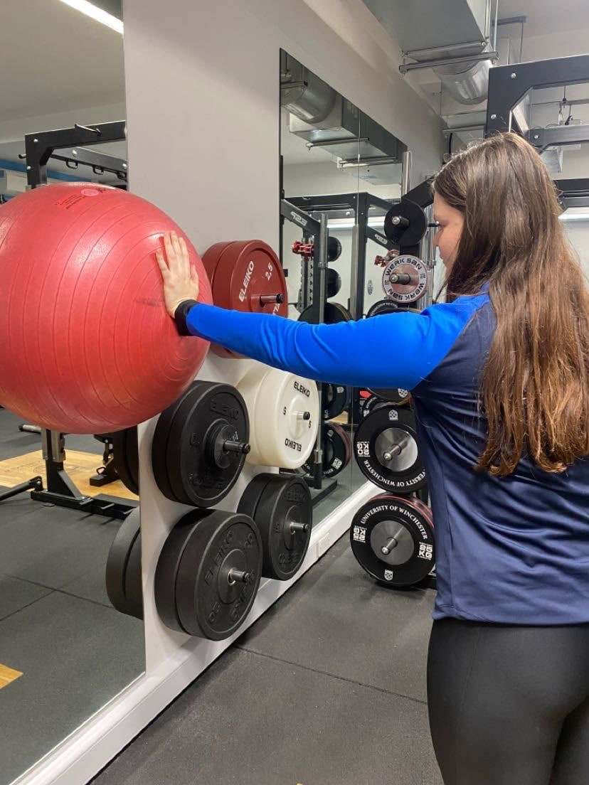 Alice demonstrating a shoulder stability exercise, pressing a gym ball into the wall